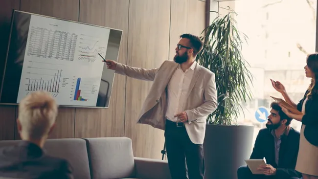 Man presenting charts on a television to a group in a conference room
