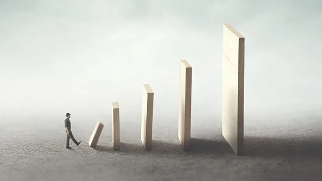 A man kicking over a small domino, which is the first in a succession of increasingly larger dominoes