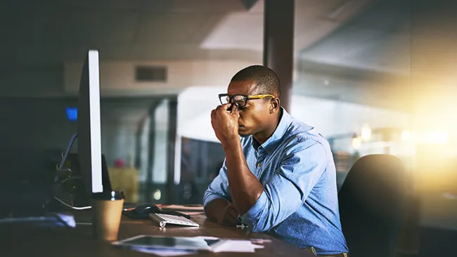 Frustrated man sitting at a computer, with this eyes closed and hands on his face