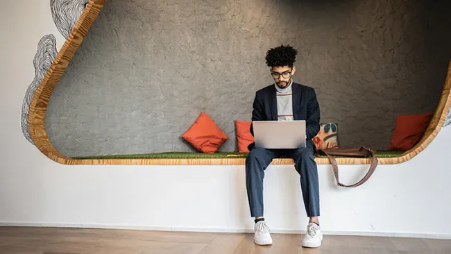 Man sitting in a modern office space, sitting on a bench with pillows and working on a laptop