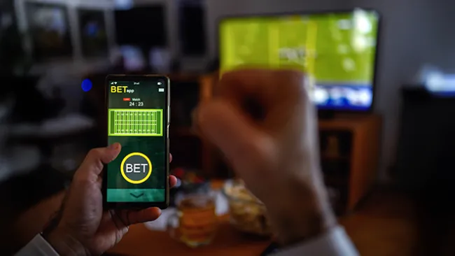 Closeup of someone's hand holding a smartphone with a sports betting app showing, with a TV screen showing football in the background