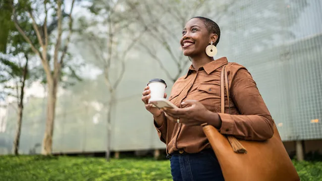 Woman smiling, holding a coffee in one hand and a smartphone in the other