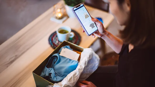 Woman looking at clothing on her phone and a piece of clothing in a box in front of her 