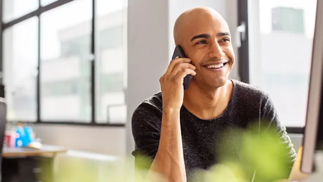 Man looking at a computer screen and holding his smartphone up to his ear, smiling