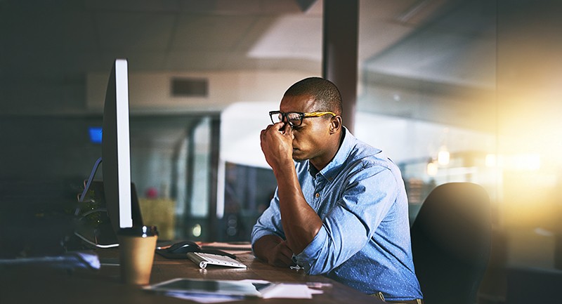 Frustrated man sitting at a computer, with this eyes closed and hands on his face