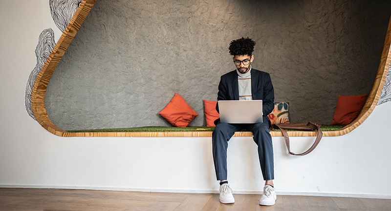 Man sitting in a modern office space, sitting on a bench with pillows and working on a laptop