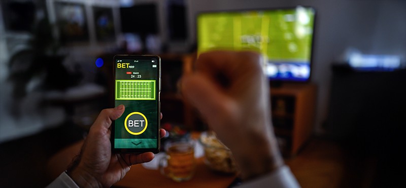 Closeup of someone's hand holding a smartphone with a sports betting app showing, with a TV screen showing football in the background