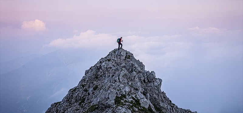 Person in hiking/climbing gear standing at the top of a mountain peak