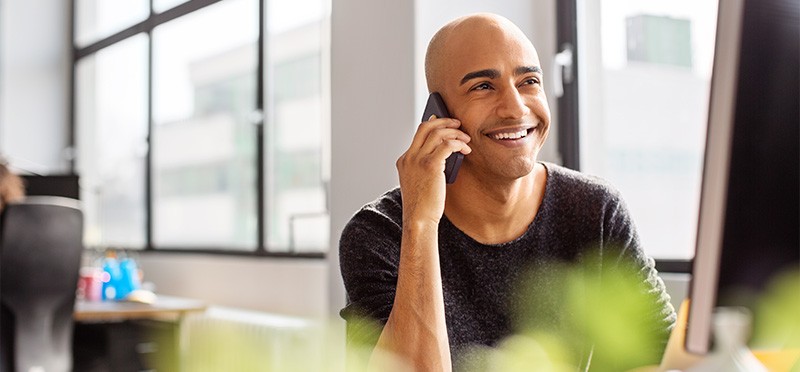 Man looking at a computer screen and holding his smartphone up to his ear, smiling