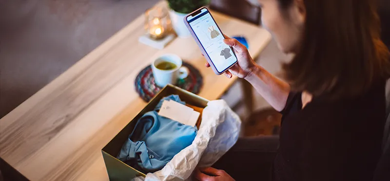 Woman looking at clothing on her phone and a piece of clothing in a box in front of her 