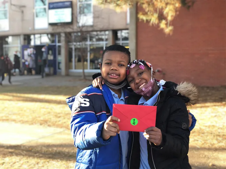 Two children holding a letter in front of a school